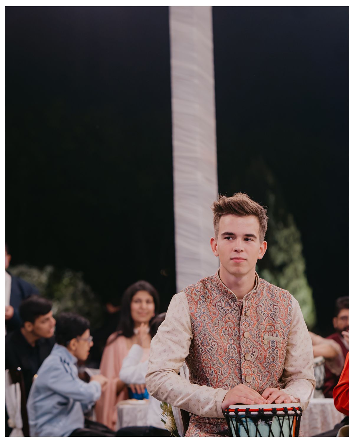 Young person in traditional Indian wedding attire playing drums at an outdoor celebration.