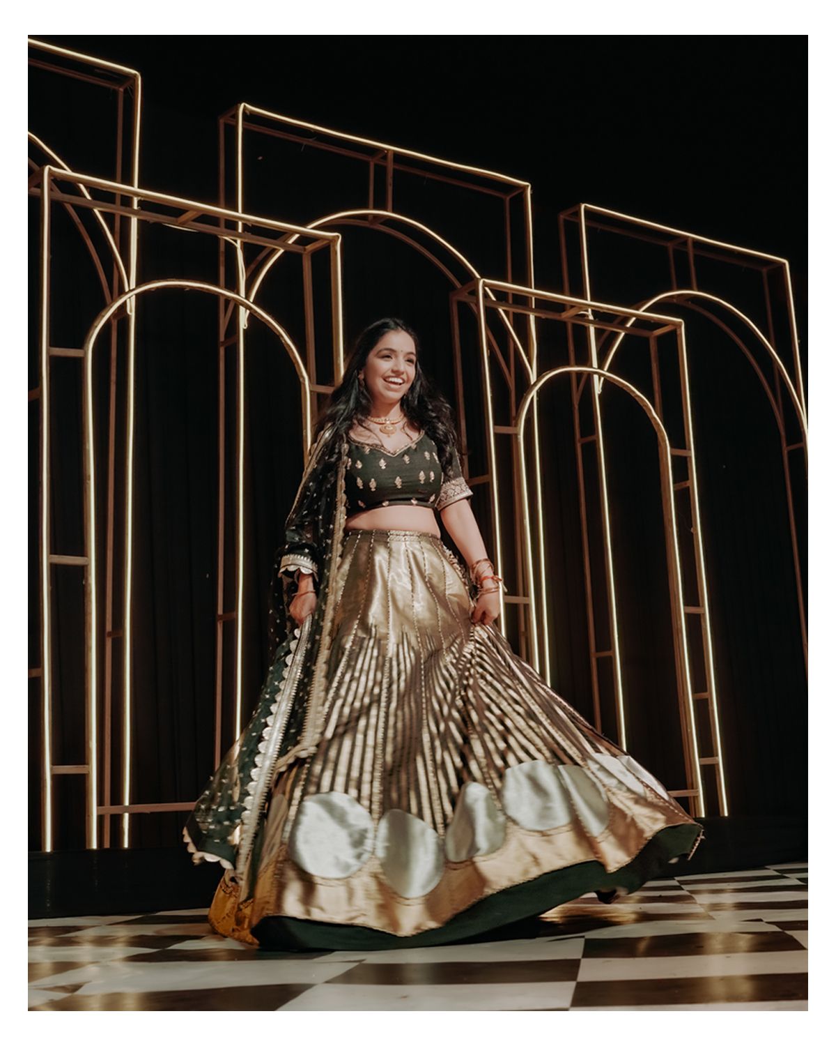 A glamorous wedding lehenga outfit in metallic gold and black twirls against an art deco geometric backdrop.