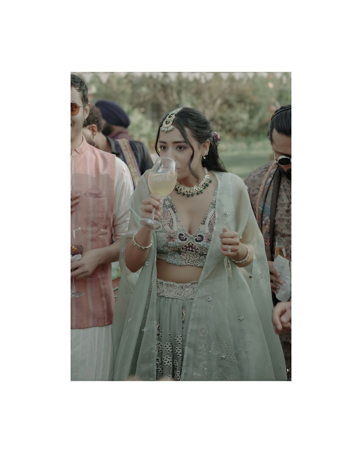 Person in an elegant mint green lehenga and crop top holding beverages at an outdoor celebration.