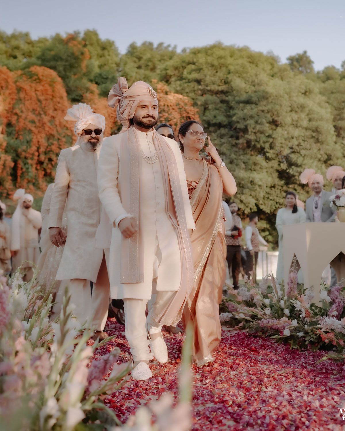 A couple walks down a flower-lined aisle covered in pink rose petals at an elegant outdoor garden wedding ceremony.