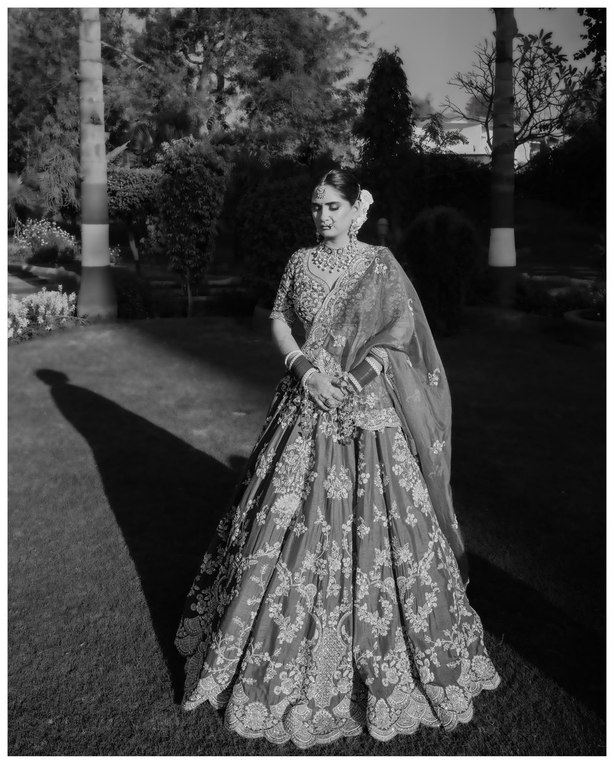Black and white portrait of a bride in an embellished lehenga standing in a garden setting.