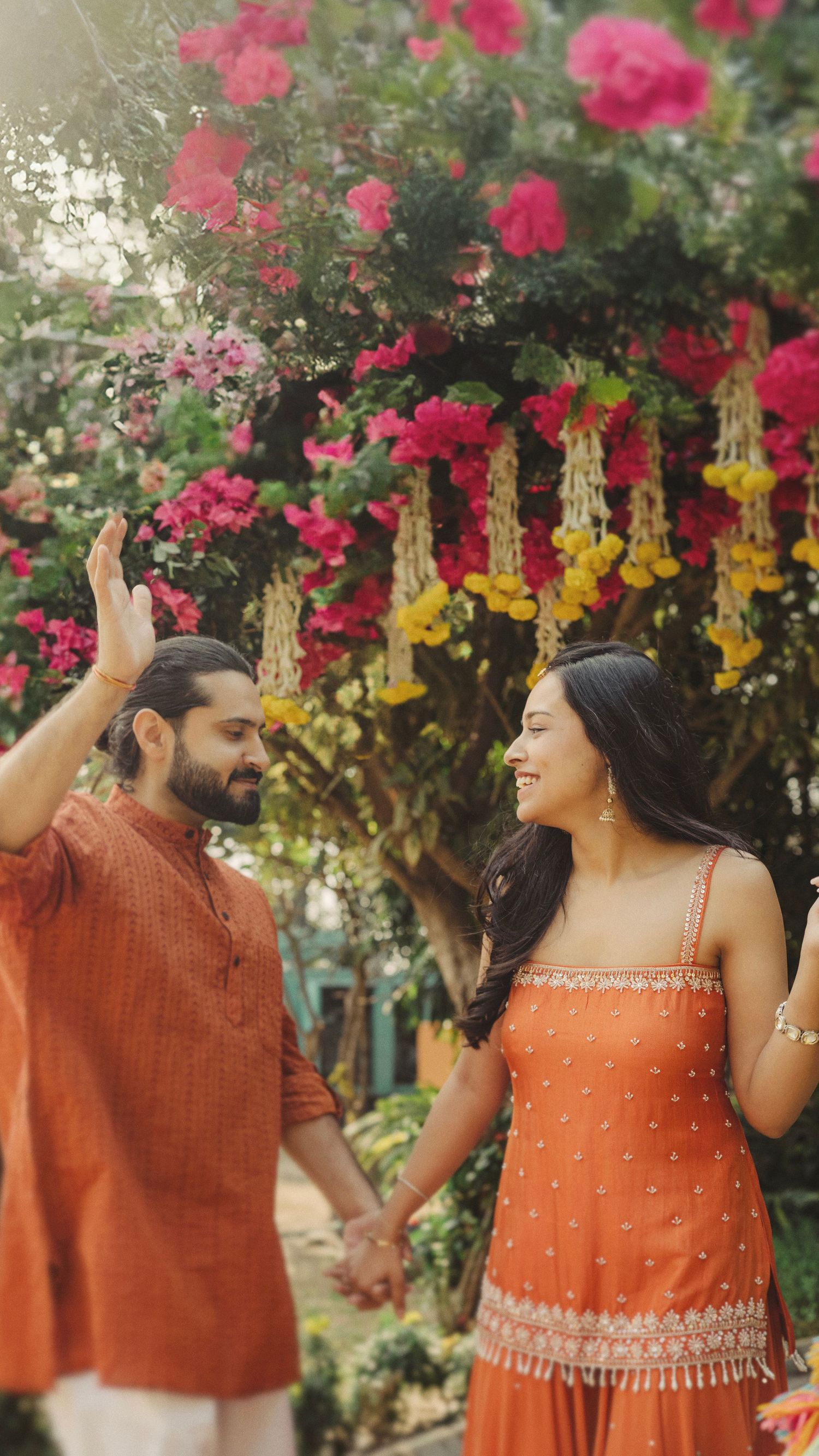 A couple in matching orange outfits dance together under pink and yellow flowers in a garden setting.