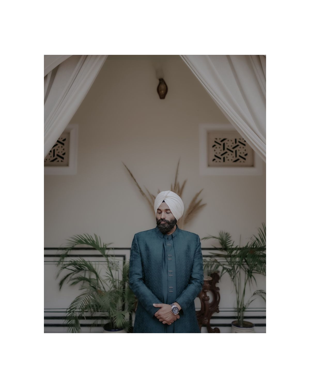A person in a teal formal suit and turban stands against a cream wall with decorative architectural details and potted plants.