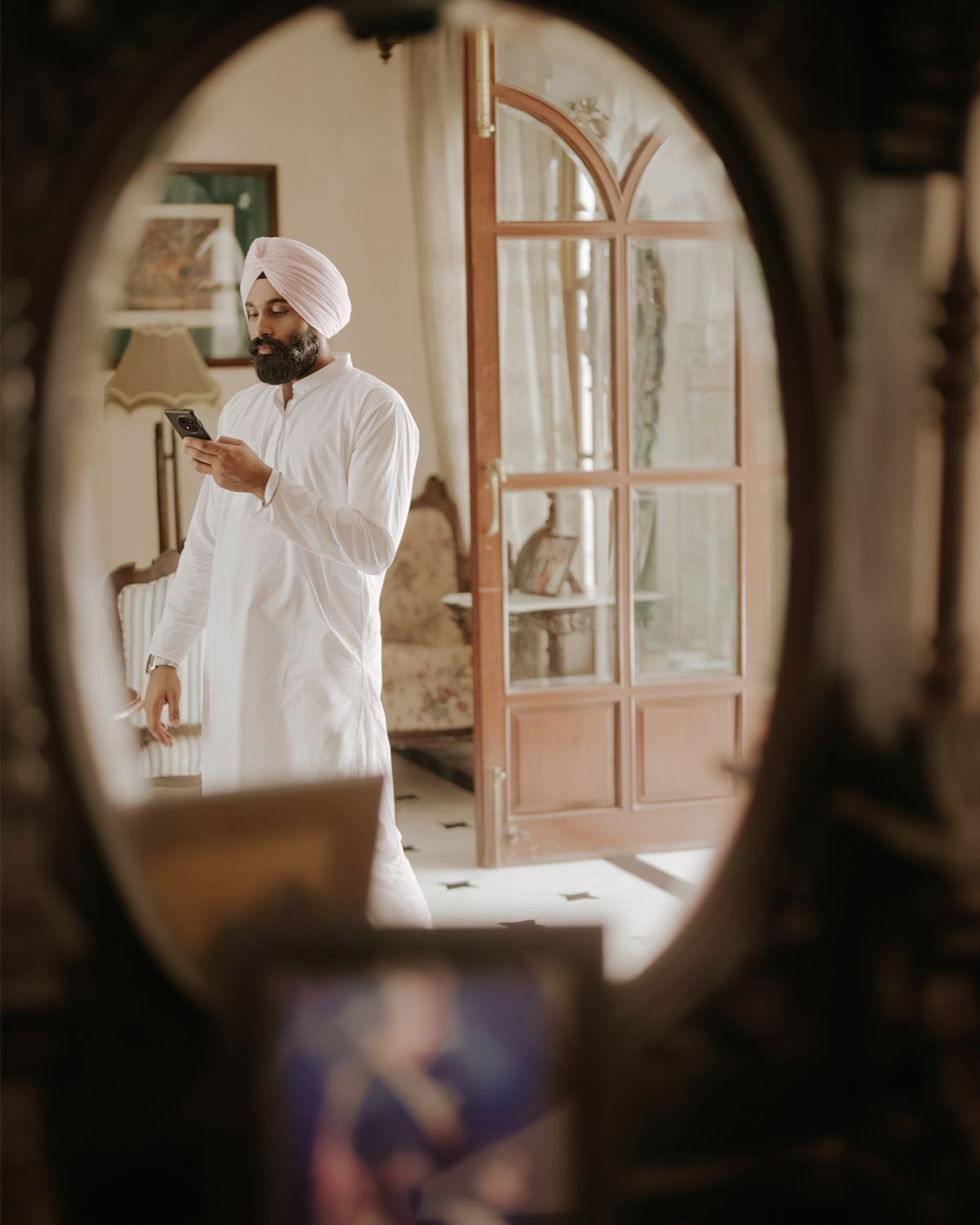 Person in white attire using phone viewed through oval mirror in elegant interior.