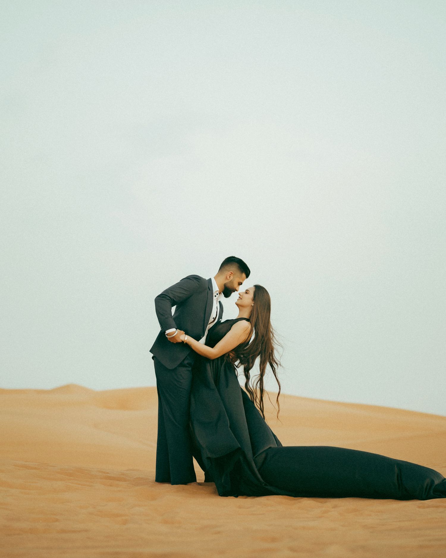 Couple in black formal attire sharing a romantic moment on golden desert sand dunes under a soft sky.