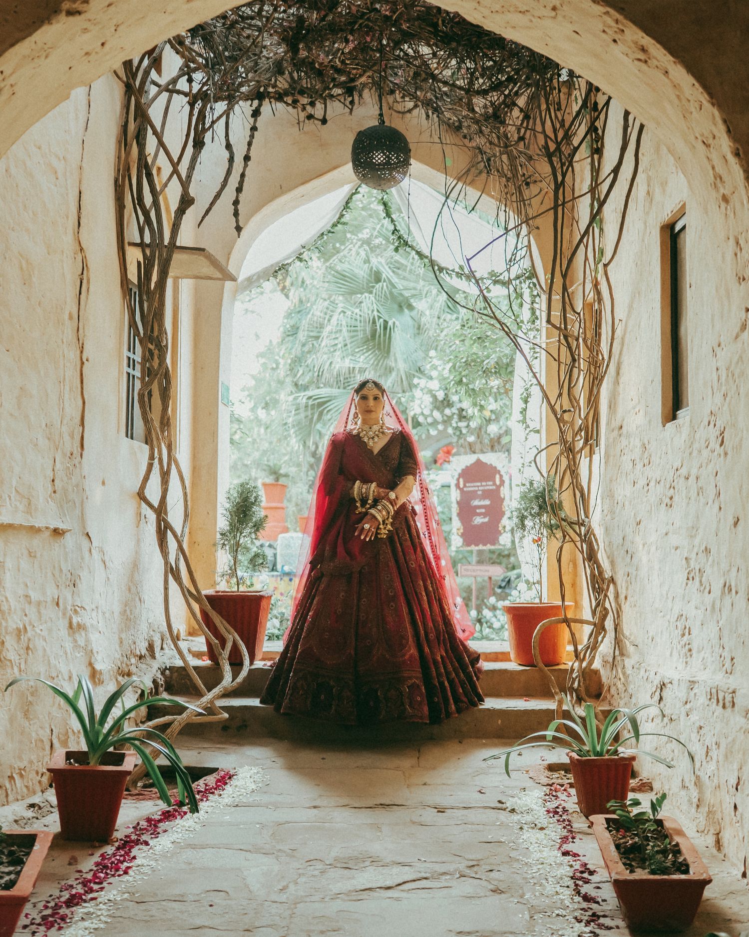 Indian bride in red lehenga standing in ornate arched doorway with vines, flower petals on ground.