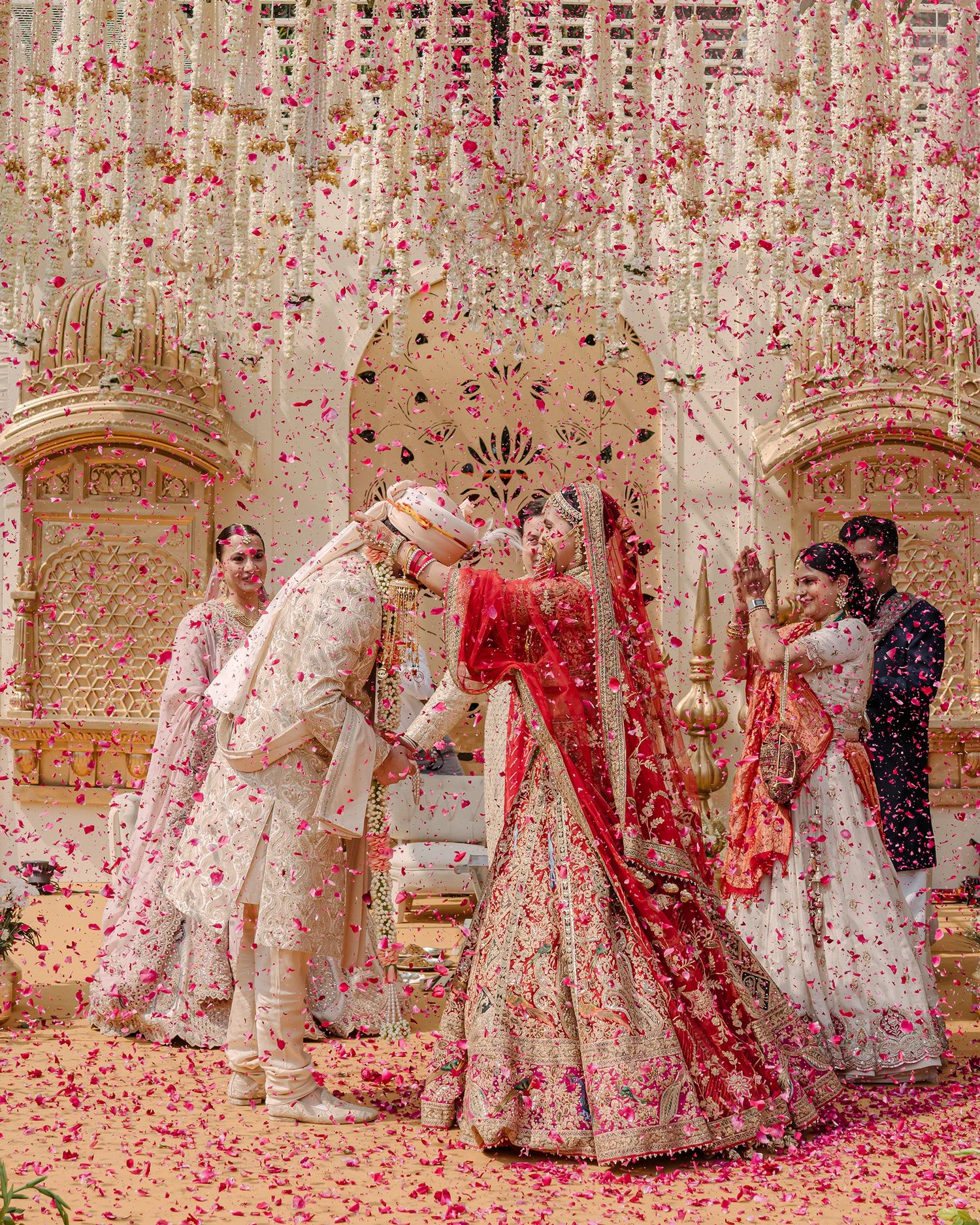 Indian bride in red lehenga and groom in white sherwani exchange vows under floral canopy with pink rose petals.