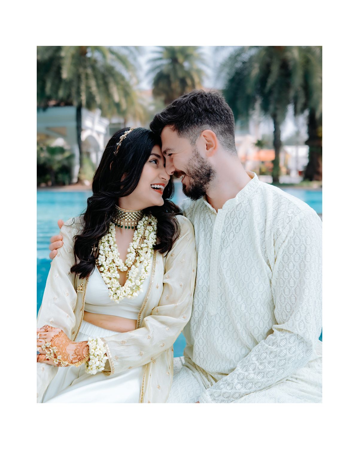 Romantic couple in white traditional outfits sharing intimate moment by pool with palm trees.