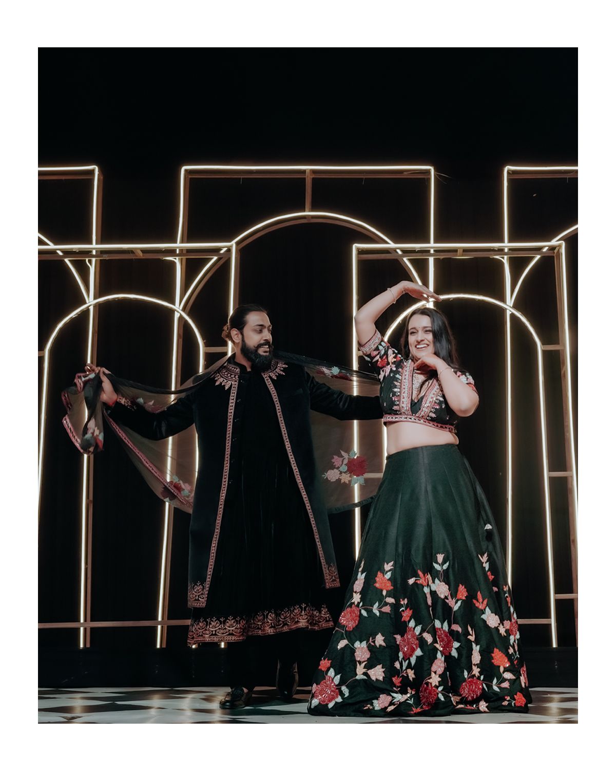 Two performers dance on stage with dramatic lighting and geometric gold archways, one wearing a floral skirt and another in dark attire.
