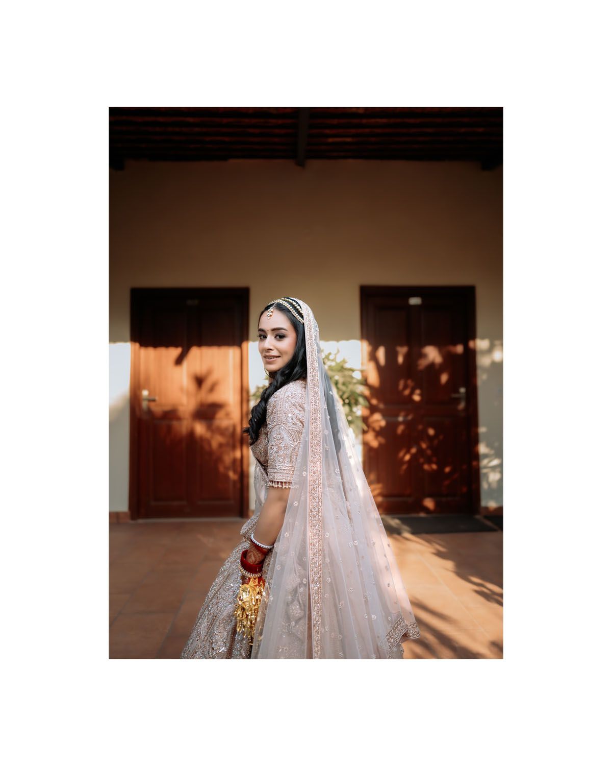 A person wearing an elegant cream-colored bridal dress with lace detailing stands in warm sunlight against a rustic backdrop.