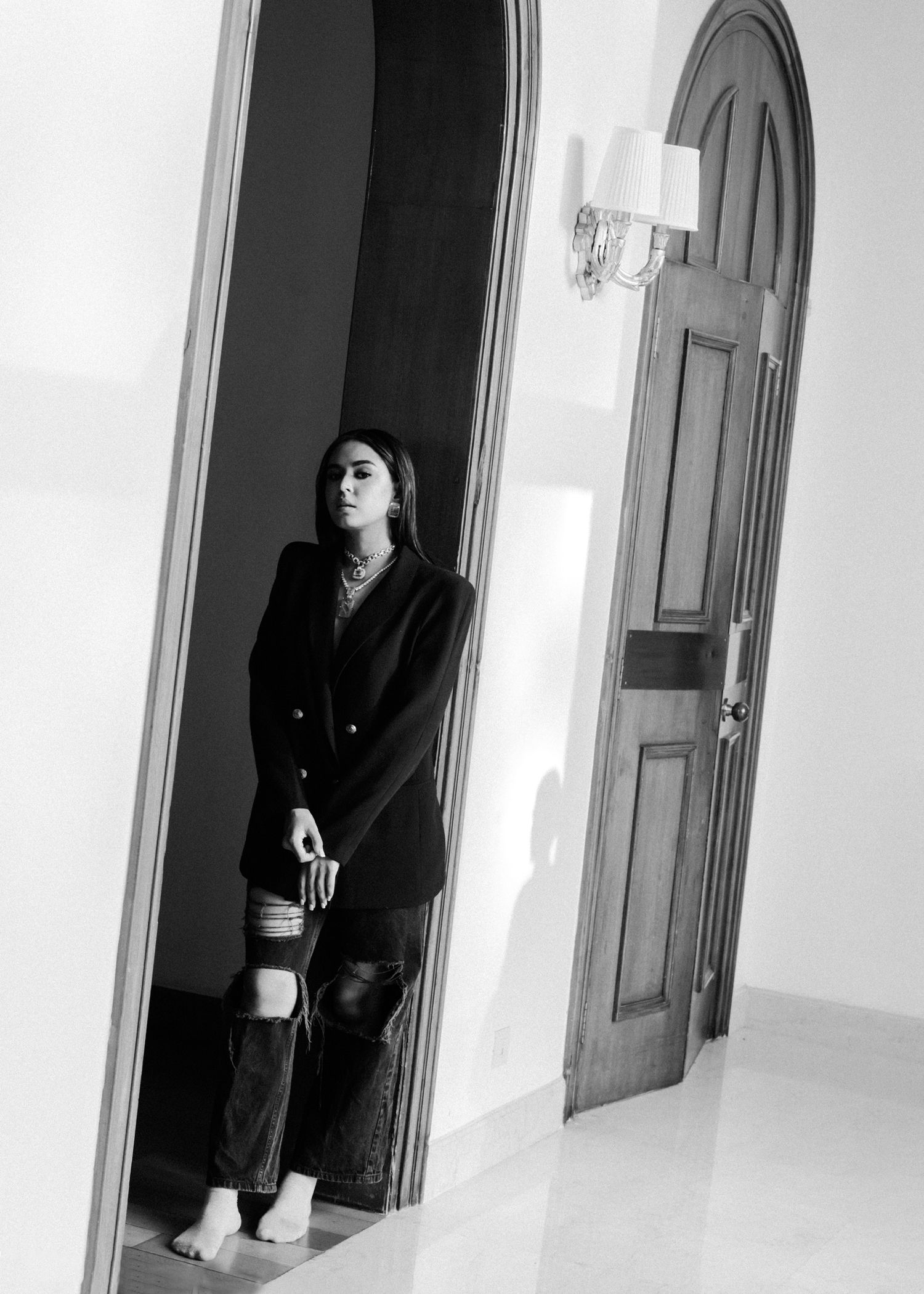 Black and white photo of a person in a dark blazer standing in an elegant hallway with arched doorways and wall sconces.