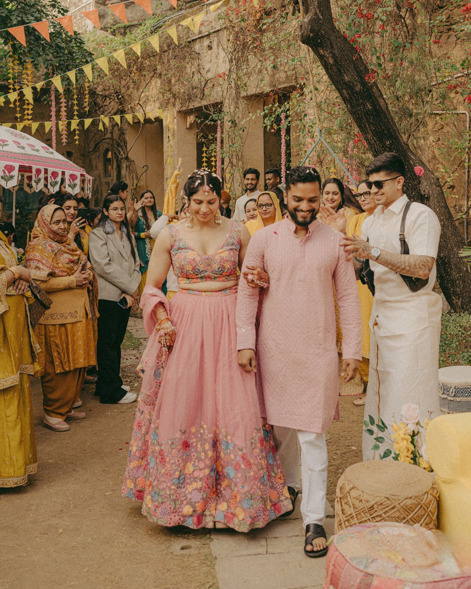Couple in matching pink outfits walk together at a vibrant Indian wedding ceremony with guests and colorful decorations.