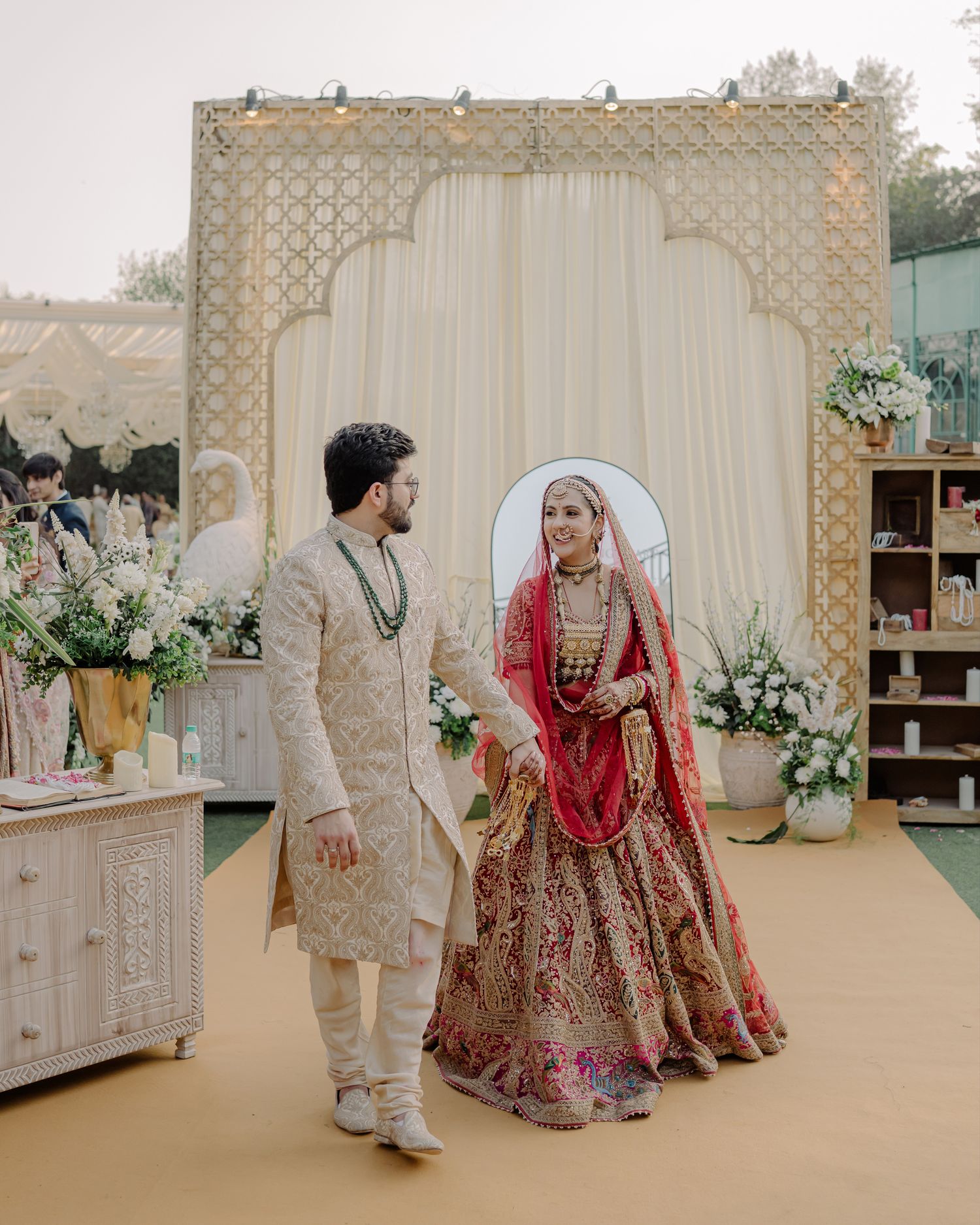 Indian bride in red and gold lehenga with groom in cream sherwani at elegant wedding ceremony backdrop.