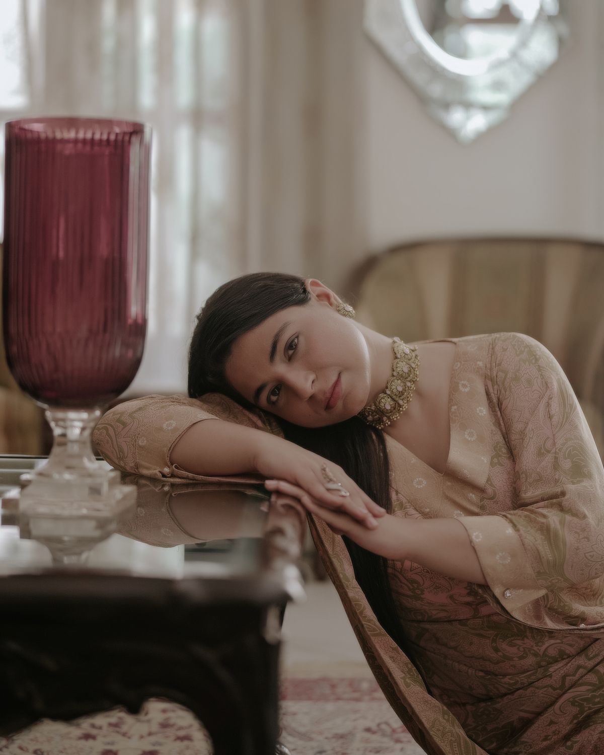 Portrait beside burgundy glass vase showing person in beige dress resting on antique furniture.