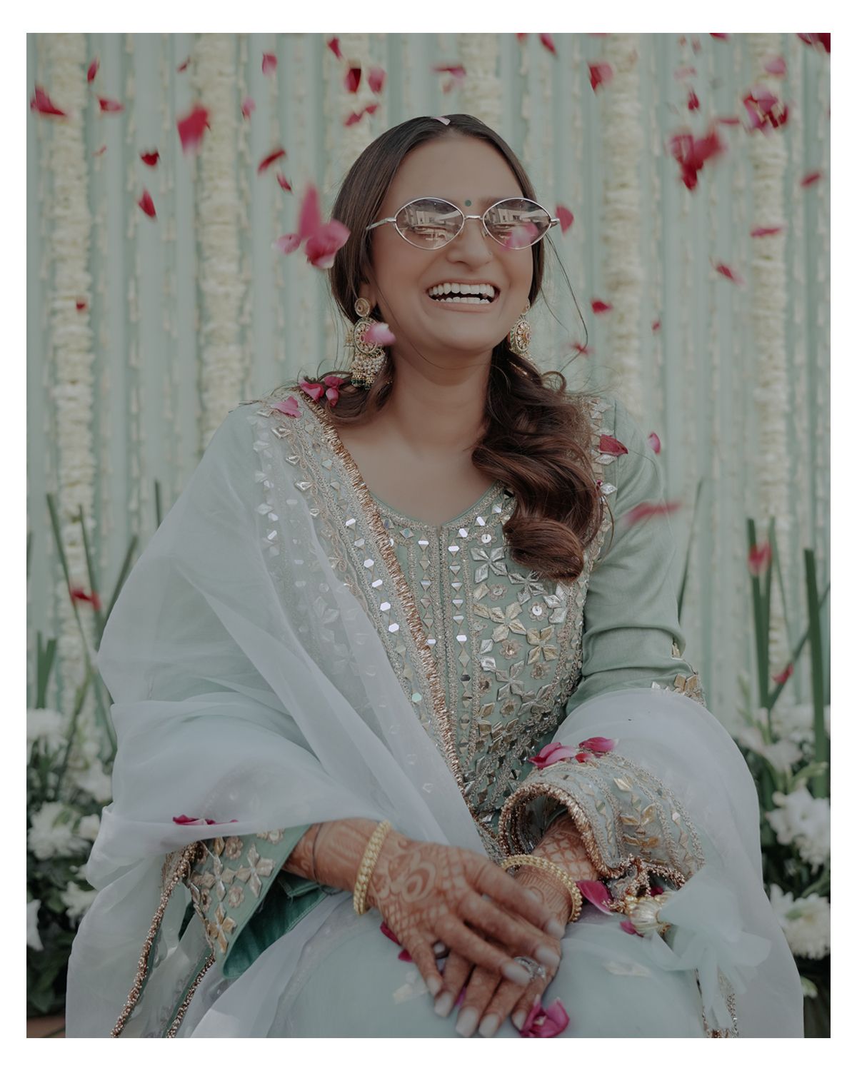 Candid portrait with rose petals falling, showing radiant smile and traditional white attire.