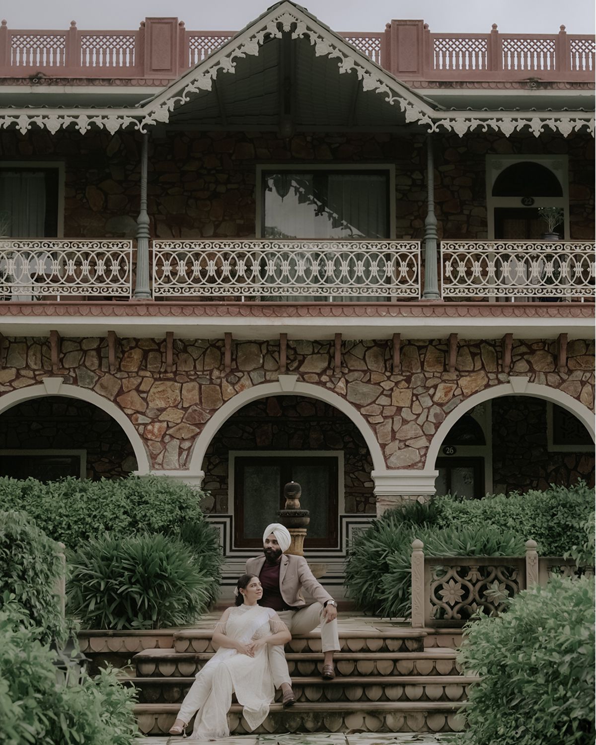 Historic stone building with ornate Victorian balcony and couple seated on entrance steps.