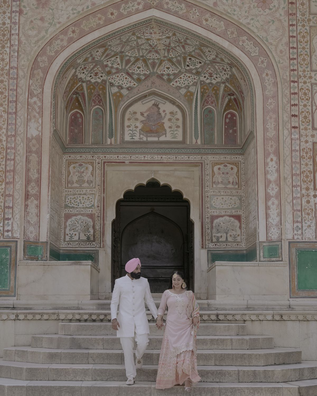 A couple standing on stone steps under an ornate Mughal-style archway with intricate wall paintings.