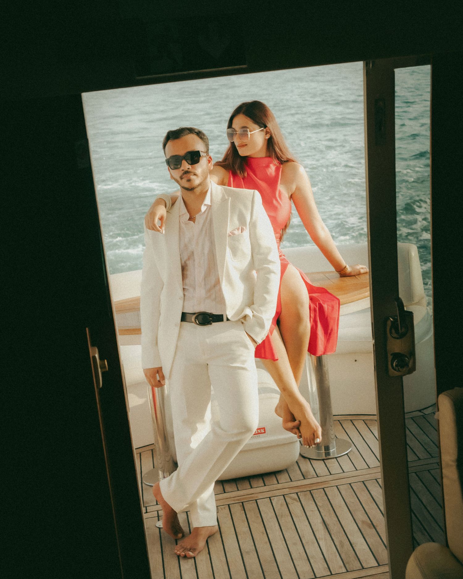Stylish couple posing on a luxury yacht, man in white suit with sunglasses, woman in red dress, ocean backdrop.