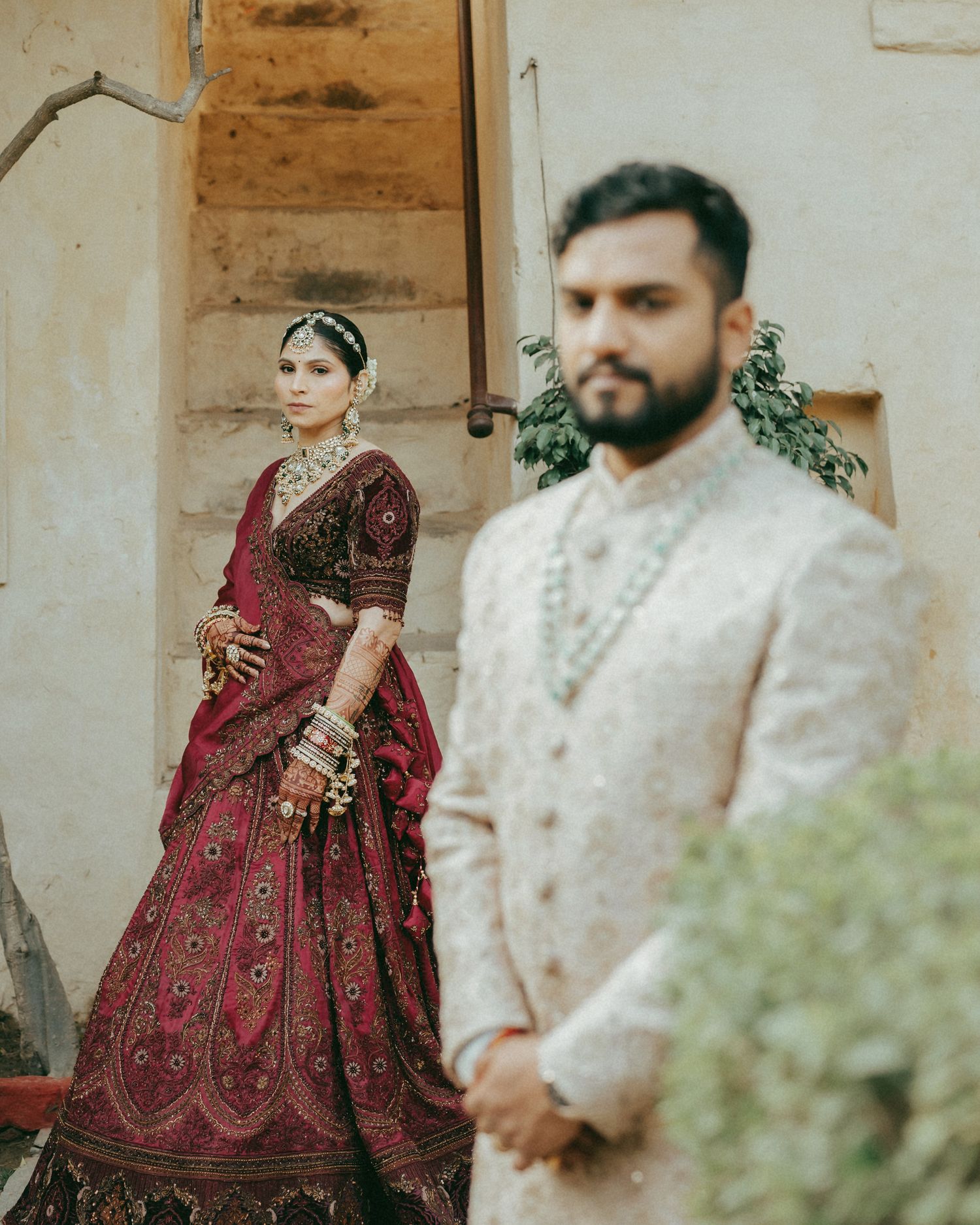 Indian bride in red lehenga and groom in ivory sherwani pose on stone steps during traditional wedding ceremony.