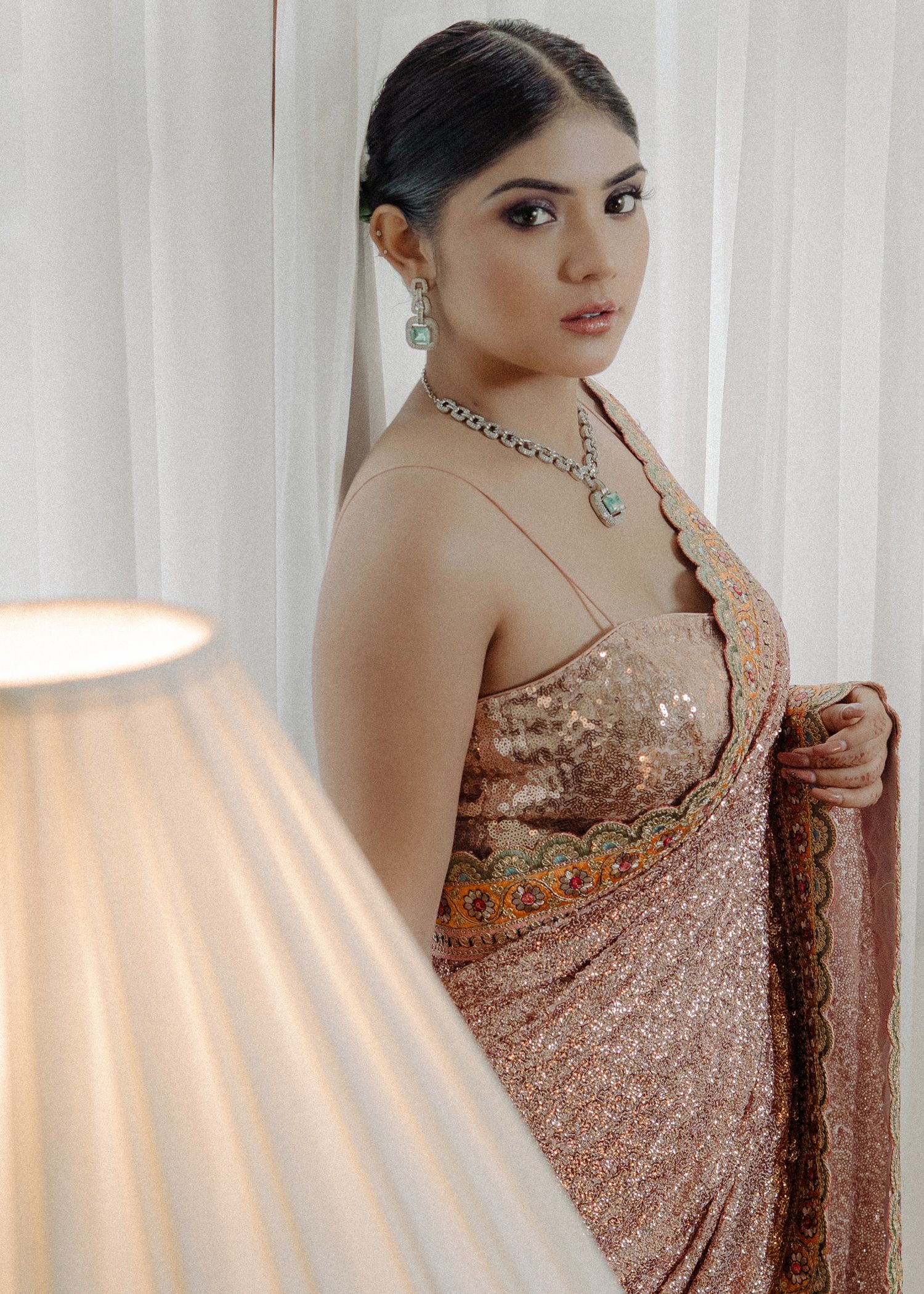Woman in sparkly rose gold sari and elegant jewelry poses against white curtain backdrop with soft lamp lighting.