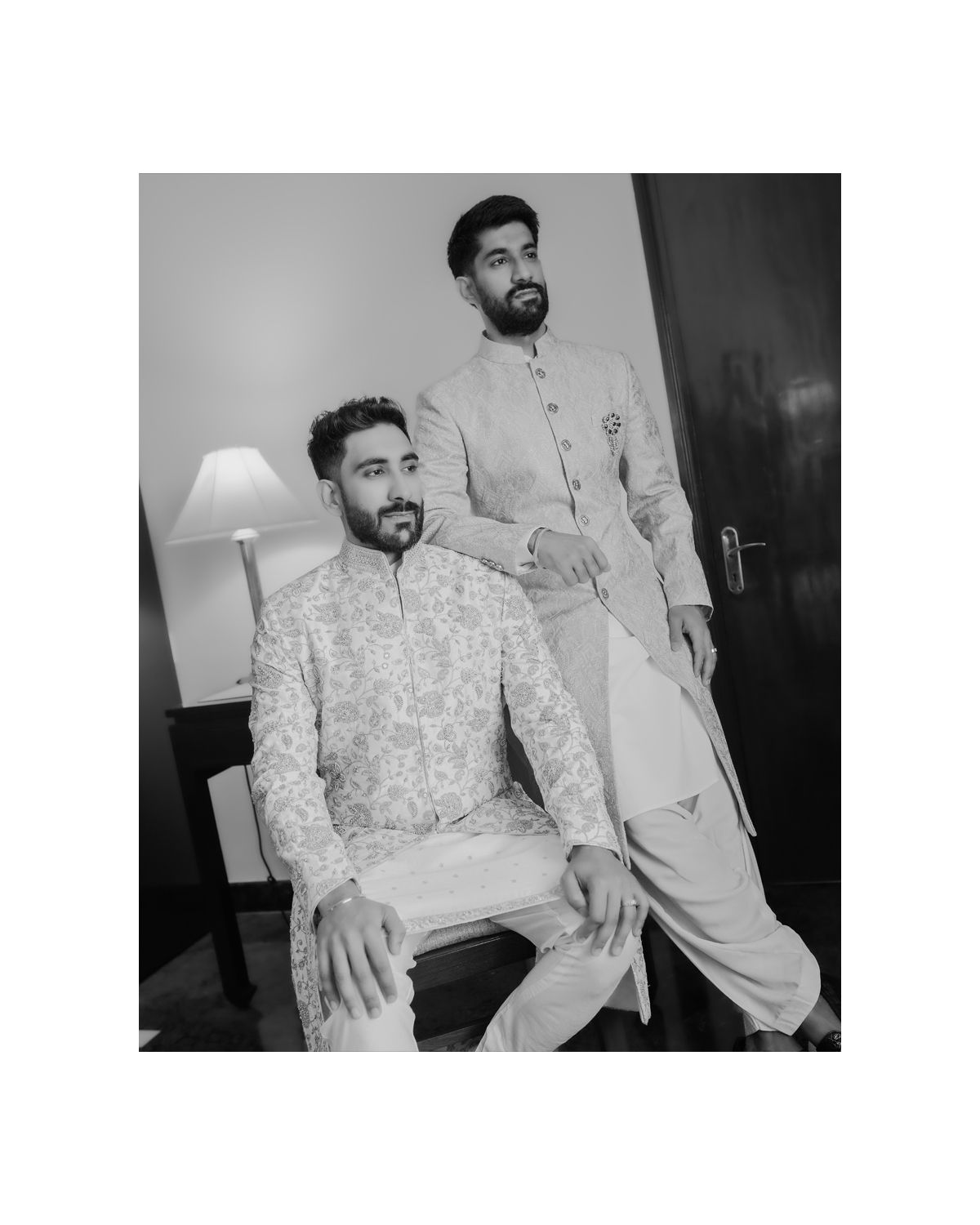 Two men in traditional white Indian formal wear pose in an indoor setting with a table lamp.