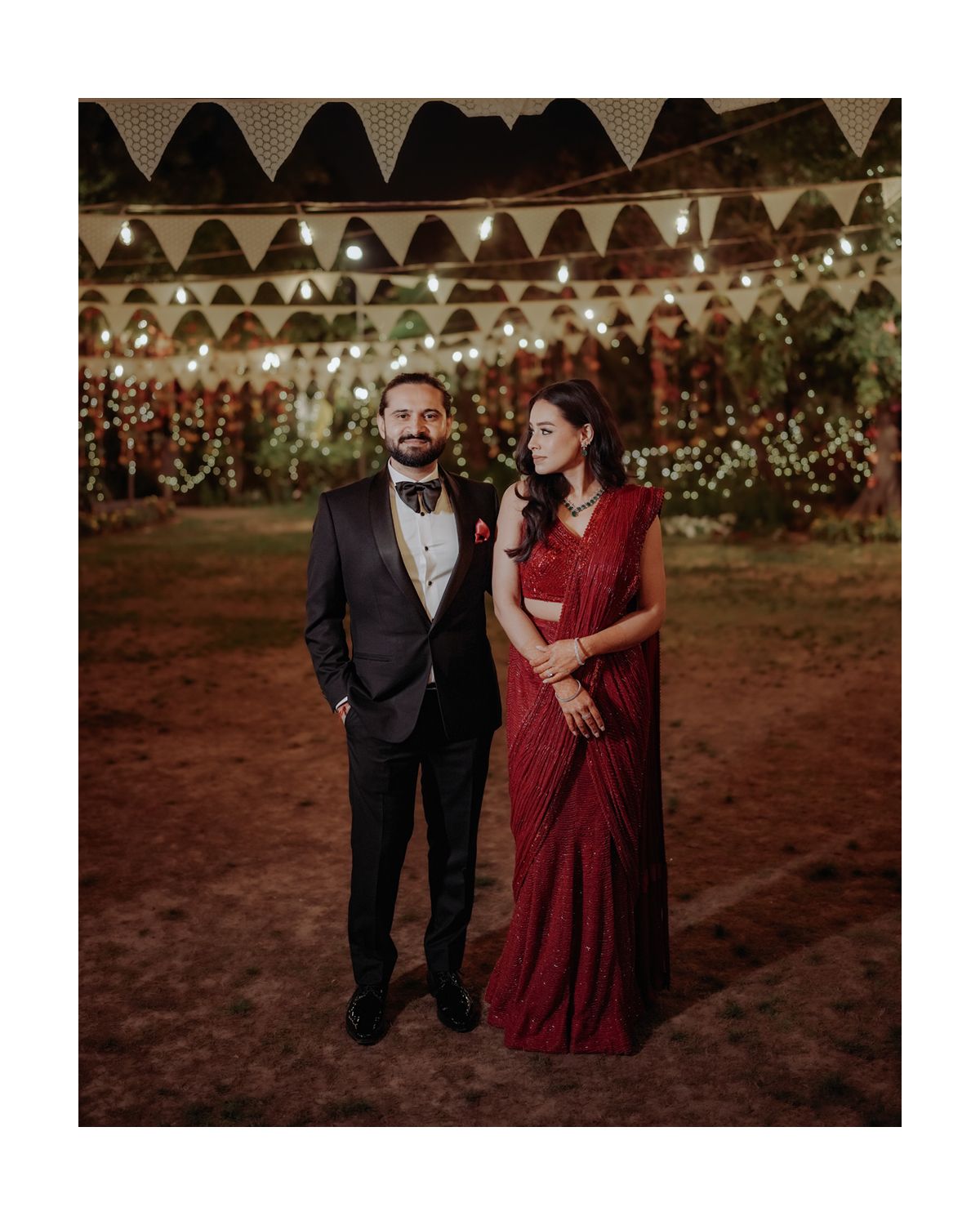 A couple in formal attire stands under decorative triangle bunting at an outdoor evening celebration.