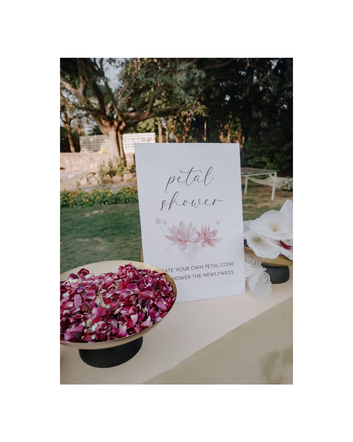 A wedding petal shower sign displayed next to a bowl of vibrant pink rose petals on a white tablecloth outdoors.