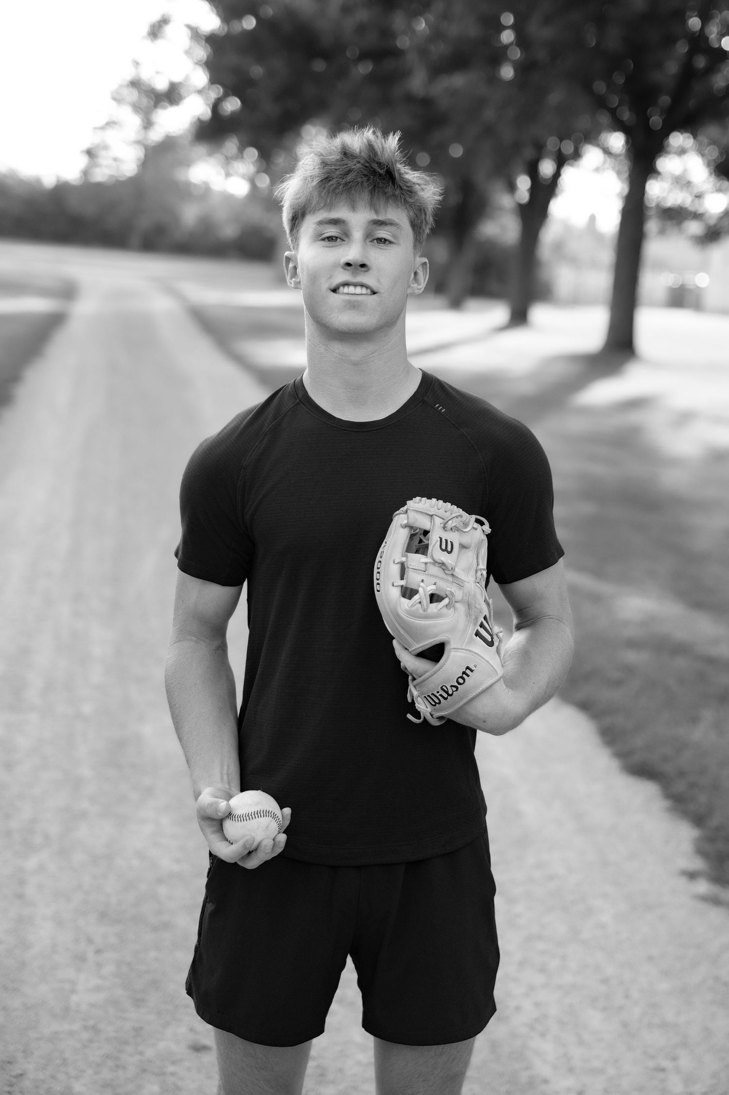 Young male baseball player holding a Wilson glove and ball on a tree-lined path, black and white photo.