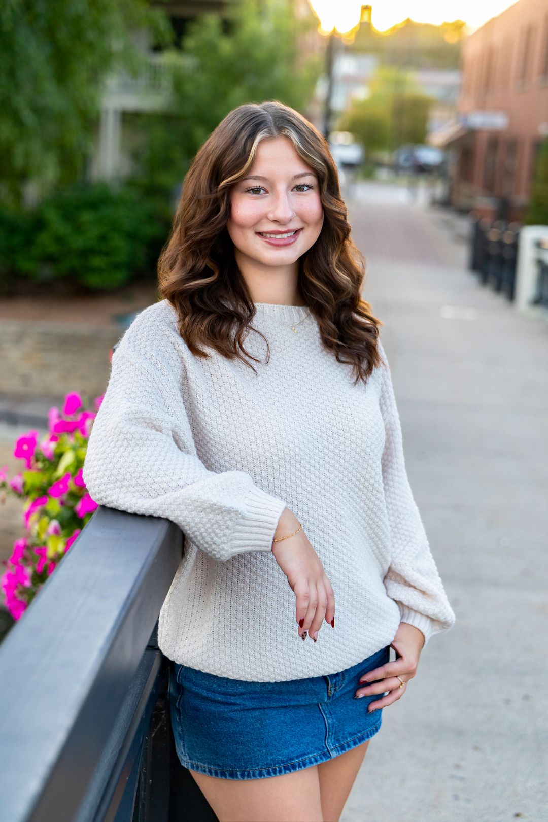 Young woman in cream sweater and denim skirt smiling outdoors on a brick walkway with pink flowers.