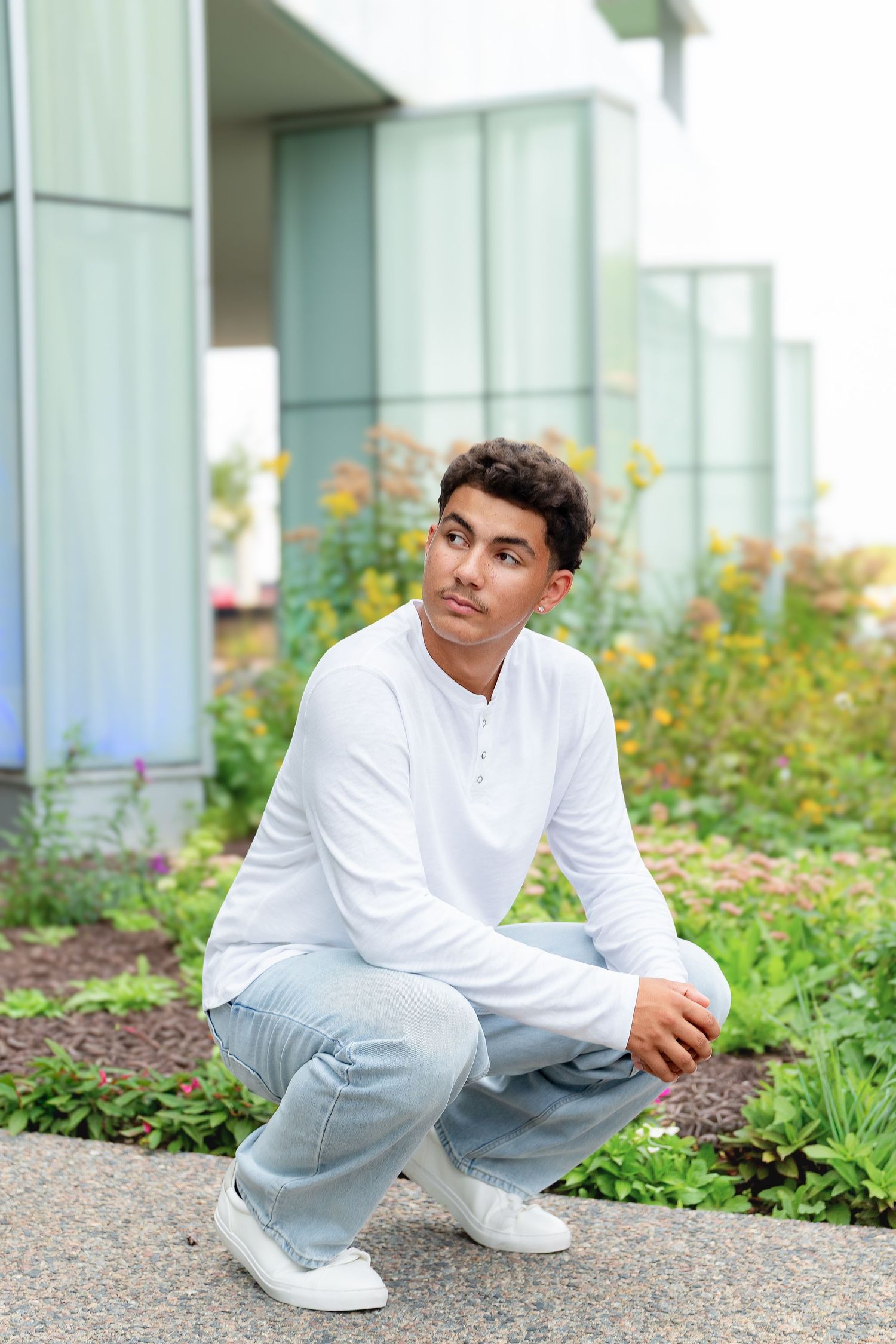 Young man in white shirt and light jeans crouching outdoors near garden with colorful flowers and modern building background.