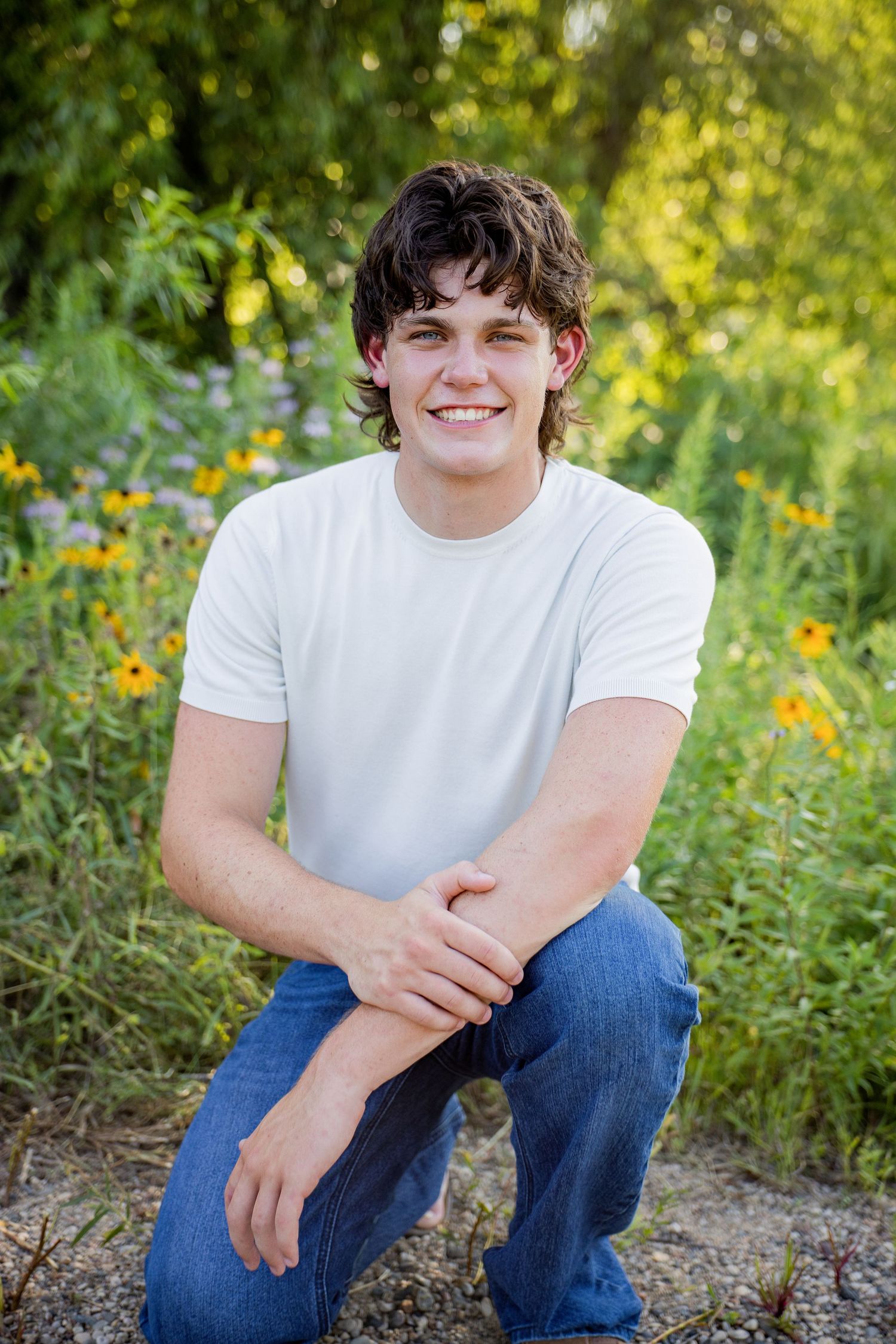 Smiling teenage boy in white t-shirt and jeans crouching outdoors among yellow wildflowers and green foliage.