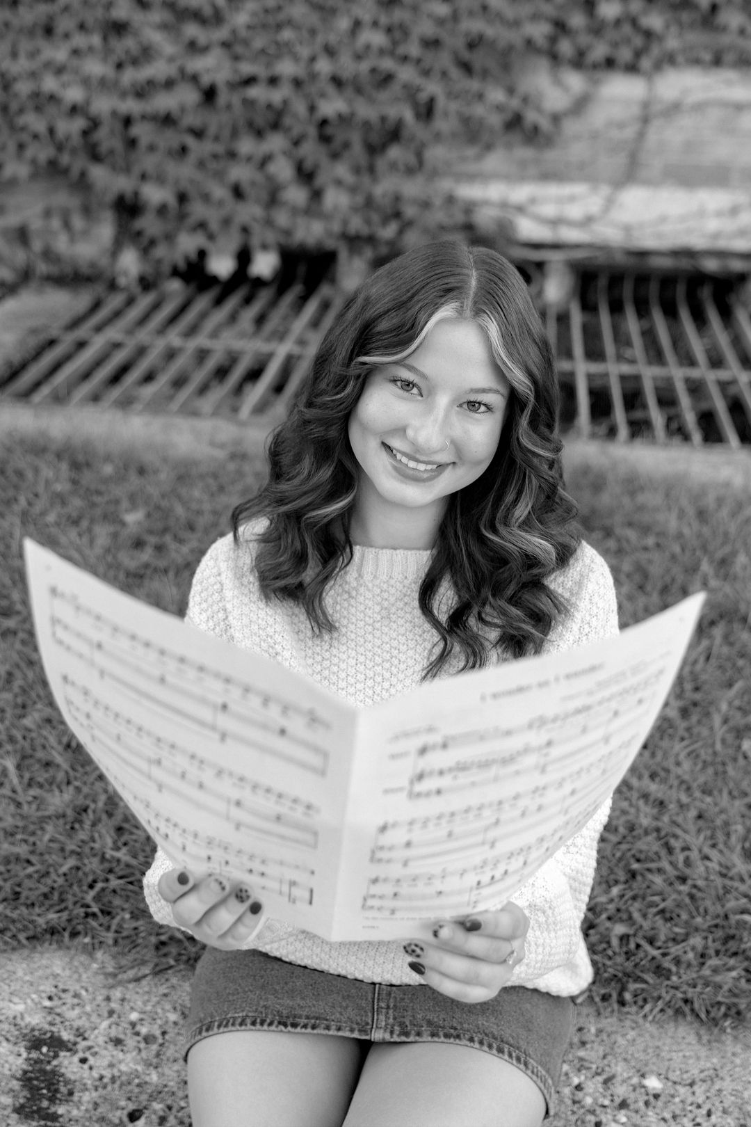 Smiling young woman holding sheet music outdoors, sitting on grass in a white sweater, black and white photo.