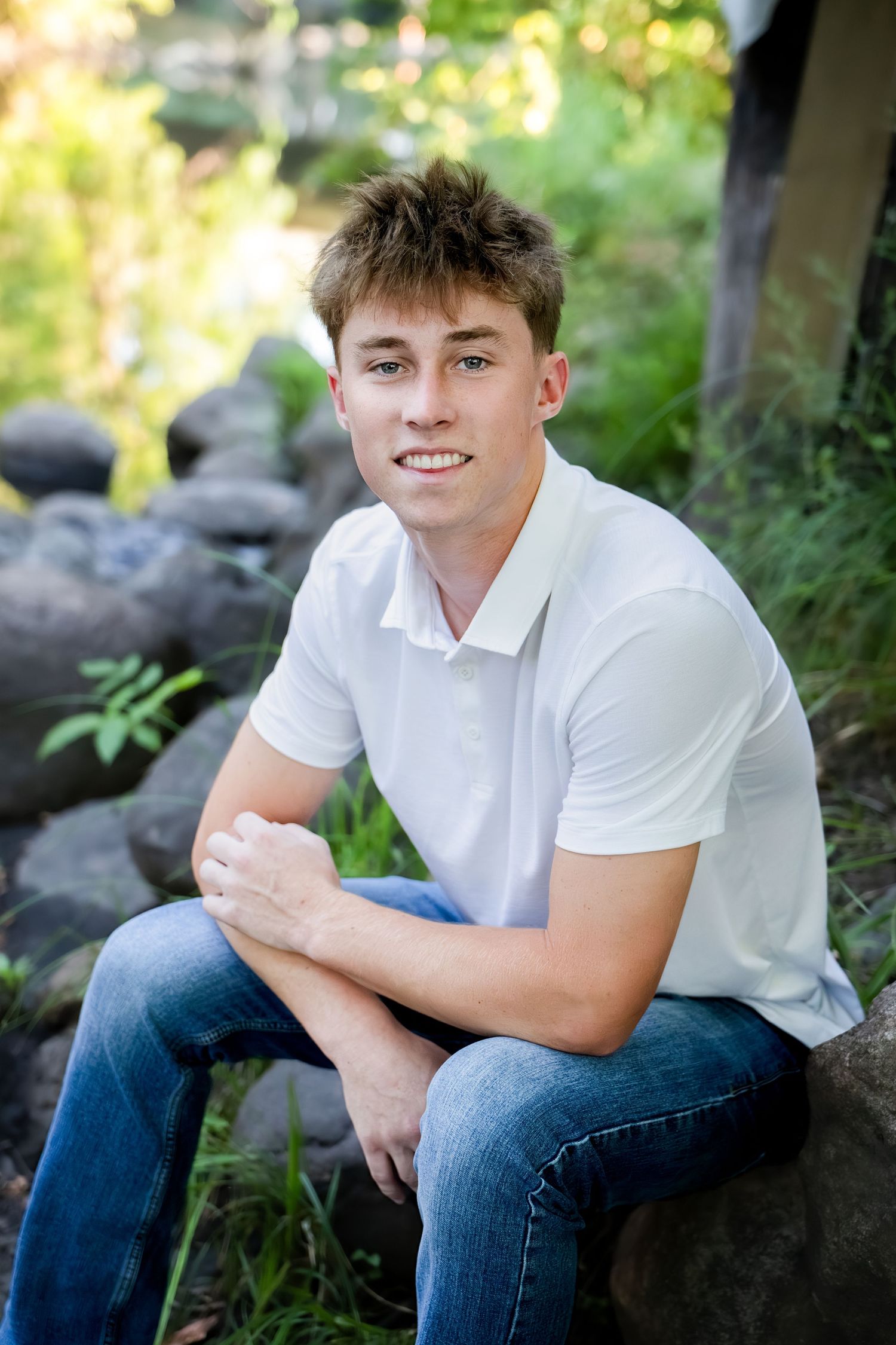 Young man in white polo shirt and jeans sitting on rocks outdoors, smiling in a lush green garden setting.