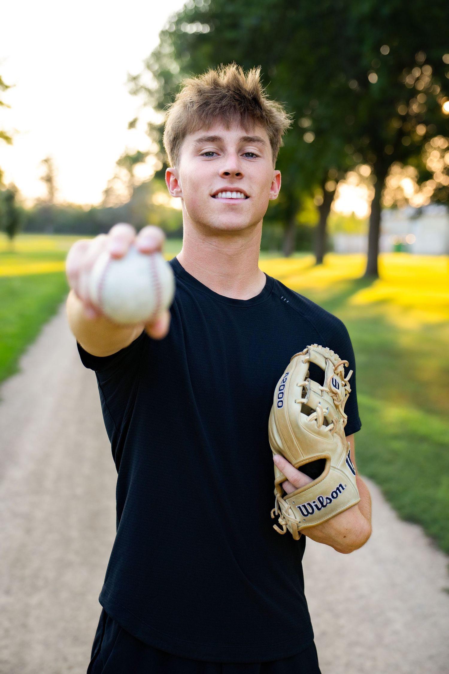 Young baseball player holding a ball toward camera with a Wilson glove outdoors in golden hour light.