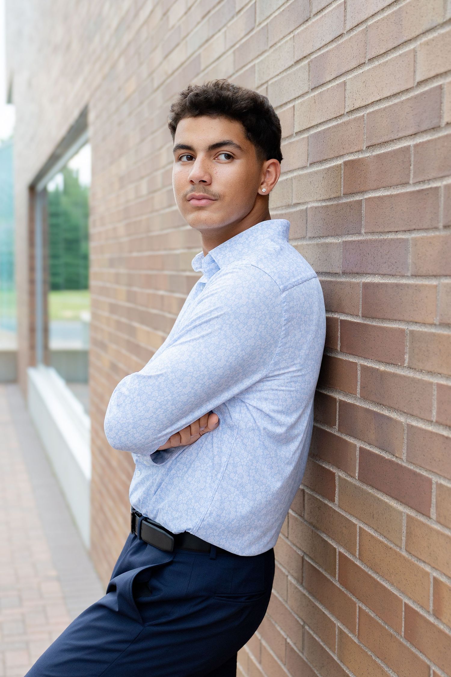Young man in light blue shirt leaning against brick wall with arms crossed, looking confidently at camera.
