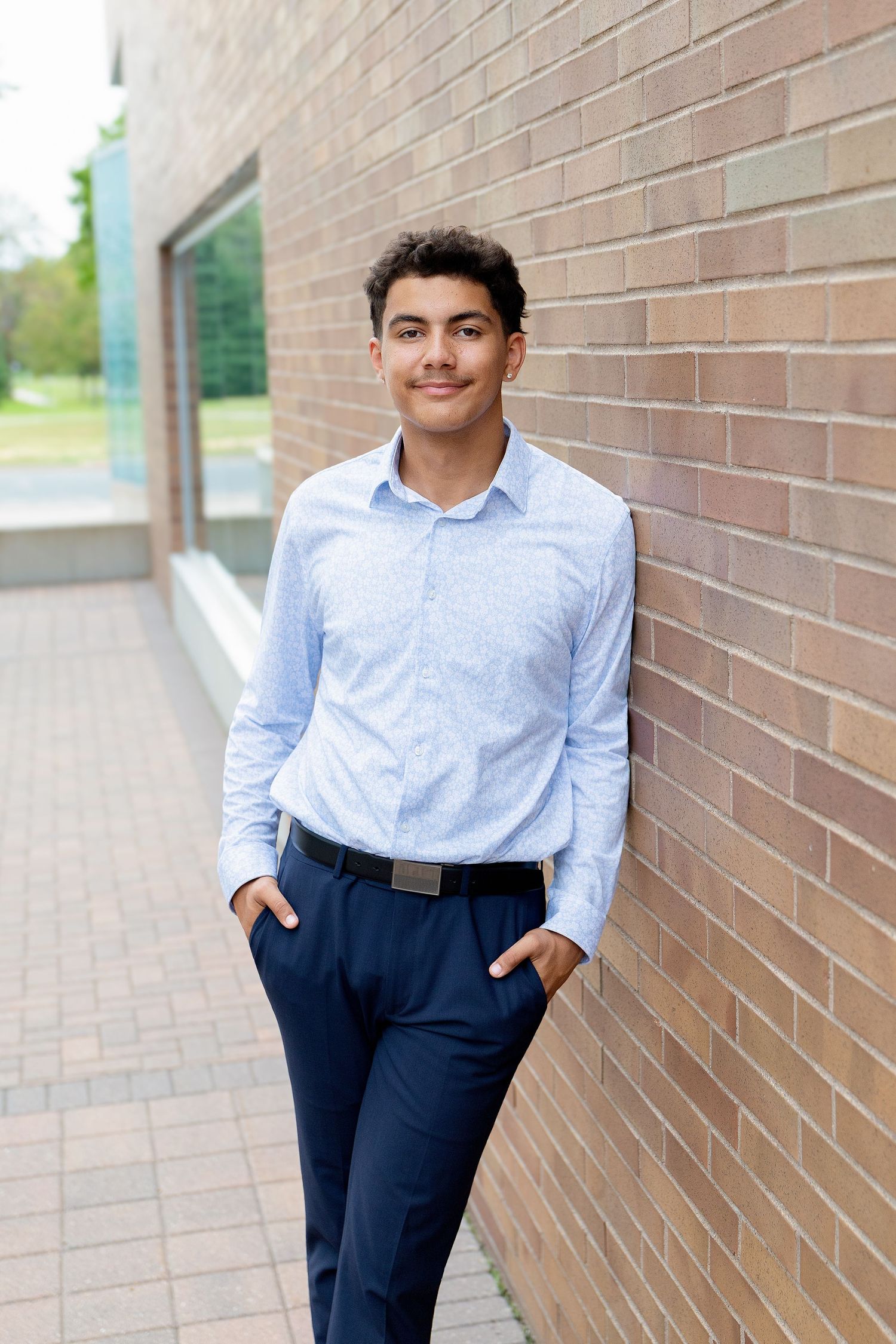 A young man in a light blue shirt and navy pants leans against a brick wall, smiling confidently outdoors.
