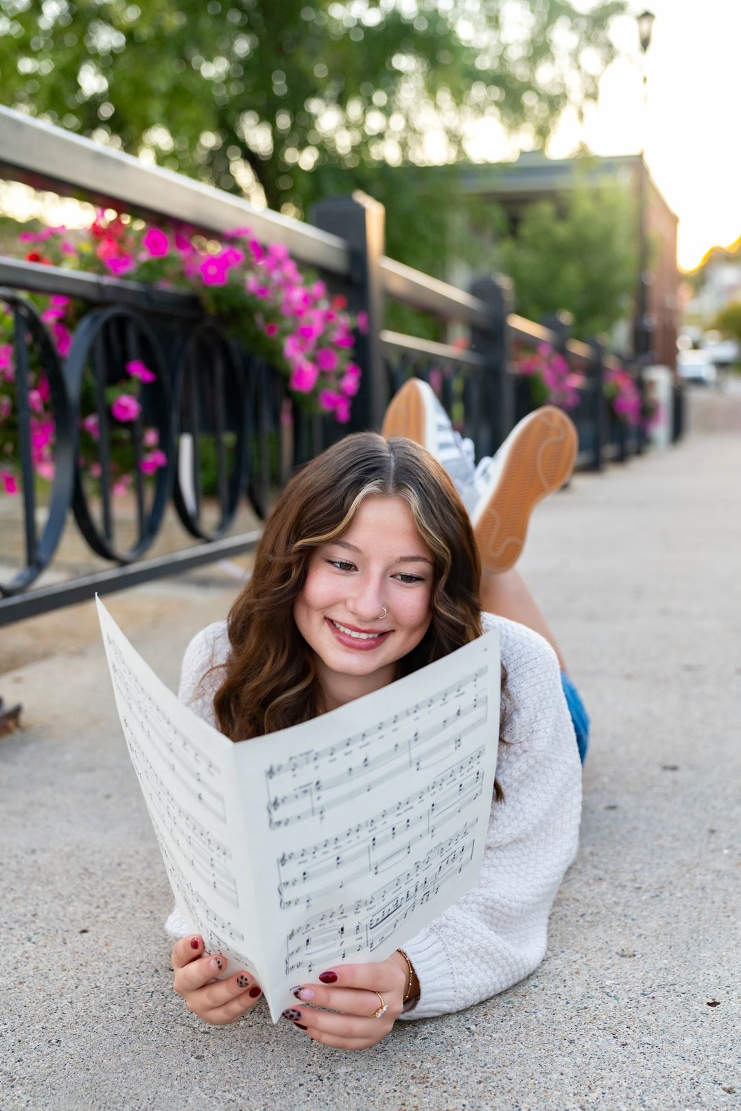 Smiling teenage girl lying on ground reading sheet music outdoors near floral railing in soft evening light.