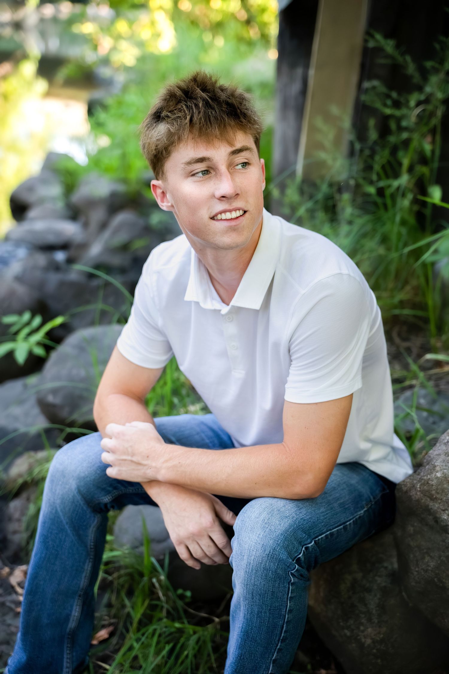 Young man in white polo shirt and jeans sitting on rocks outdoors, smiling in a lush green garden setting.
