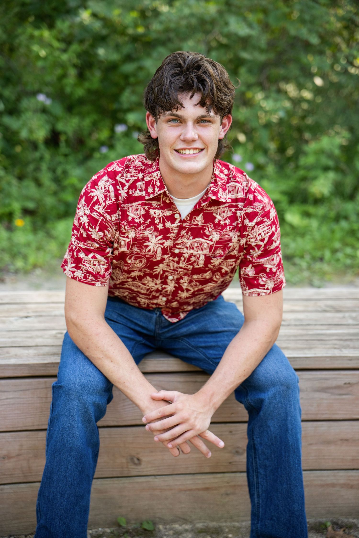 Young man in red Hawaiian shirt and jeans smiling while seated on wooden steps outdoors.