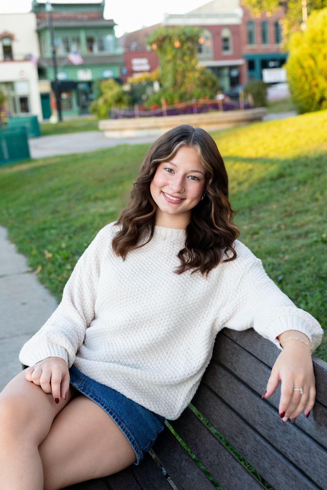 Young woman in white sweater and denim skirt smiling on a park bench with colorful storefronts behind her.