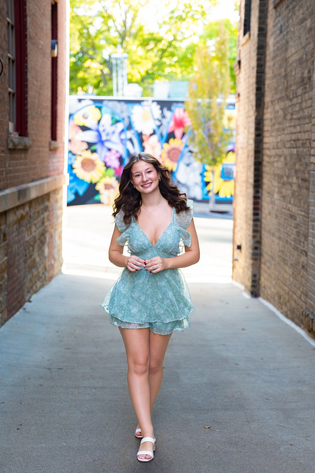 Young woman in a sage green mini dress walks through a brick alleyway with a colorful floral mural backdrop.