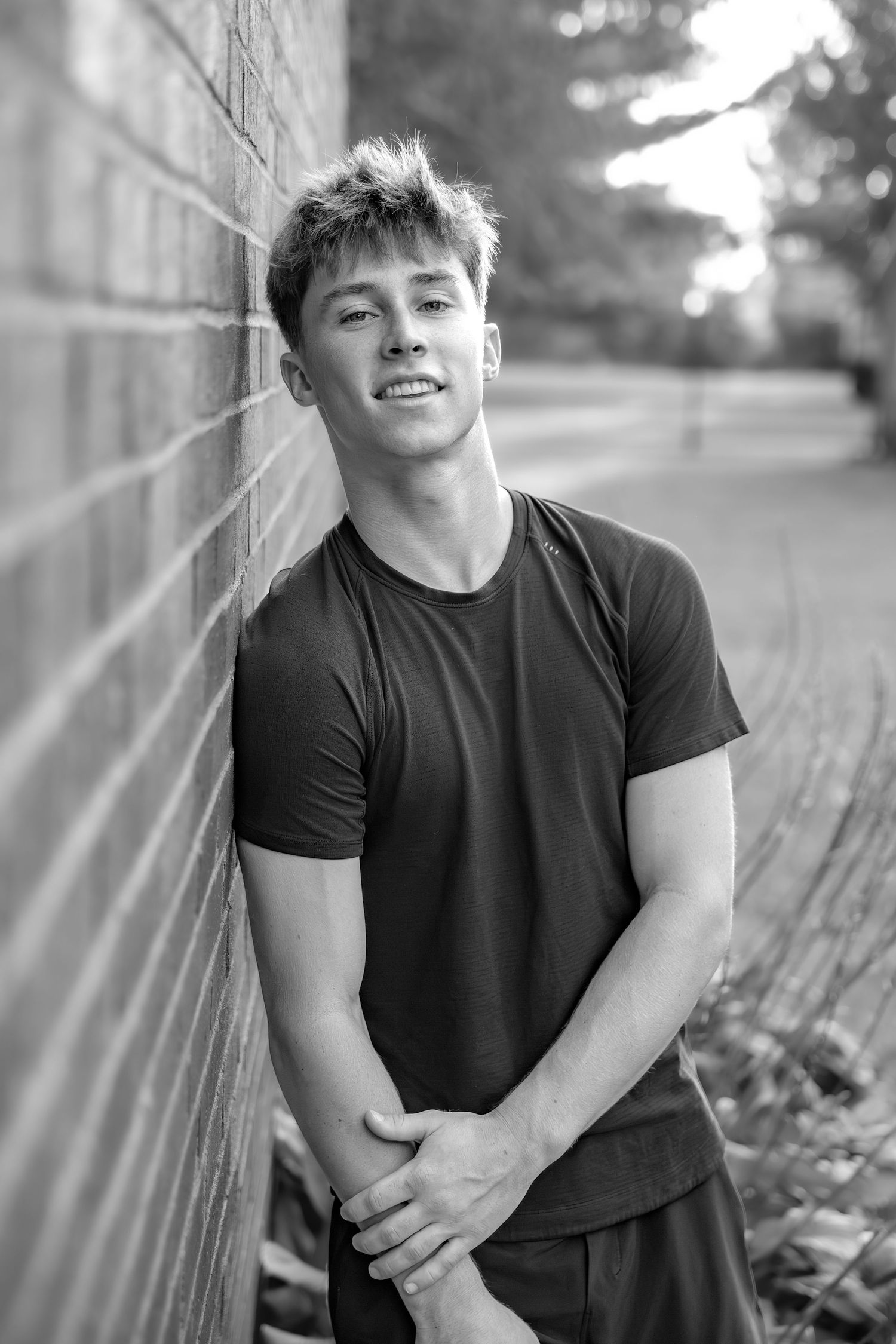 Young man in black t-shirt leaning against brick wall, smiling confidently in black and white portrait photo.