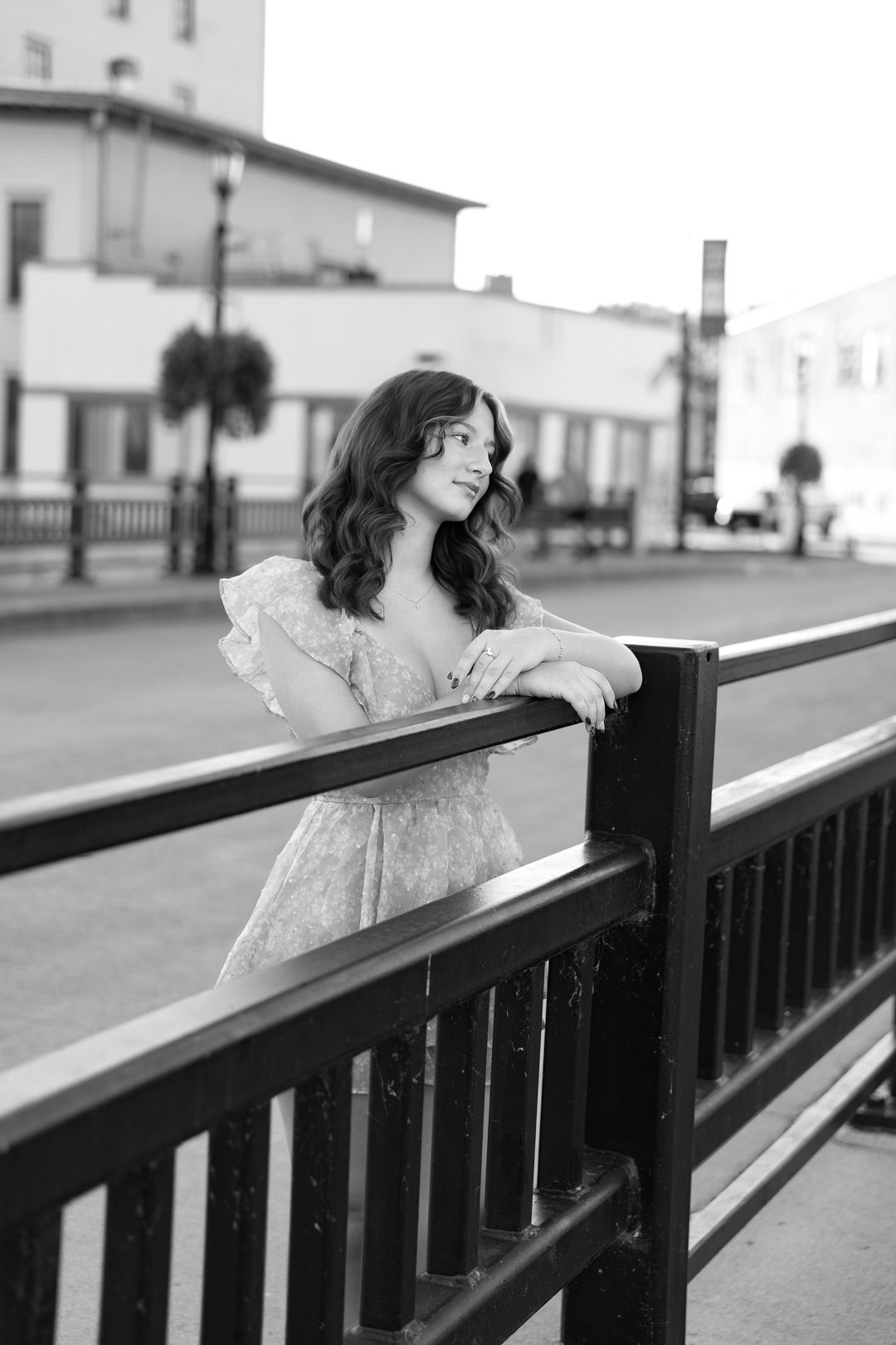 Black and white photo of a young woman in a floral dress leaning on a railing in an urban setting.
