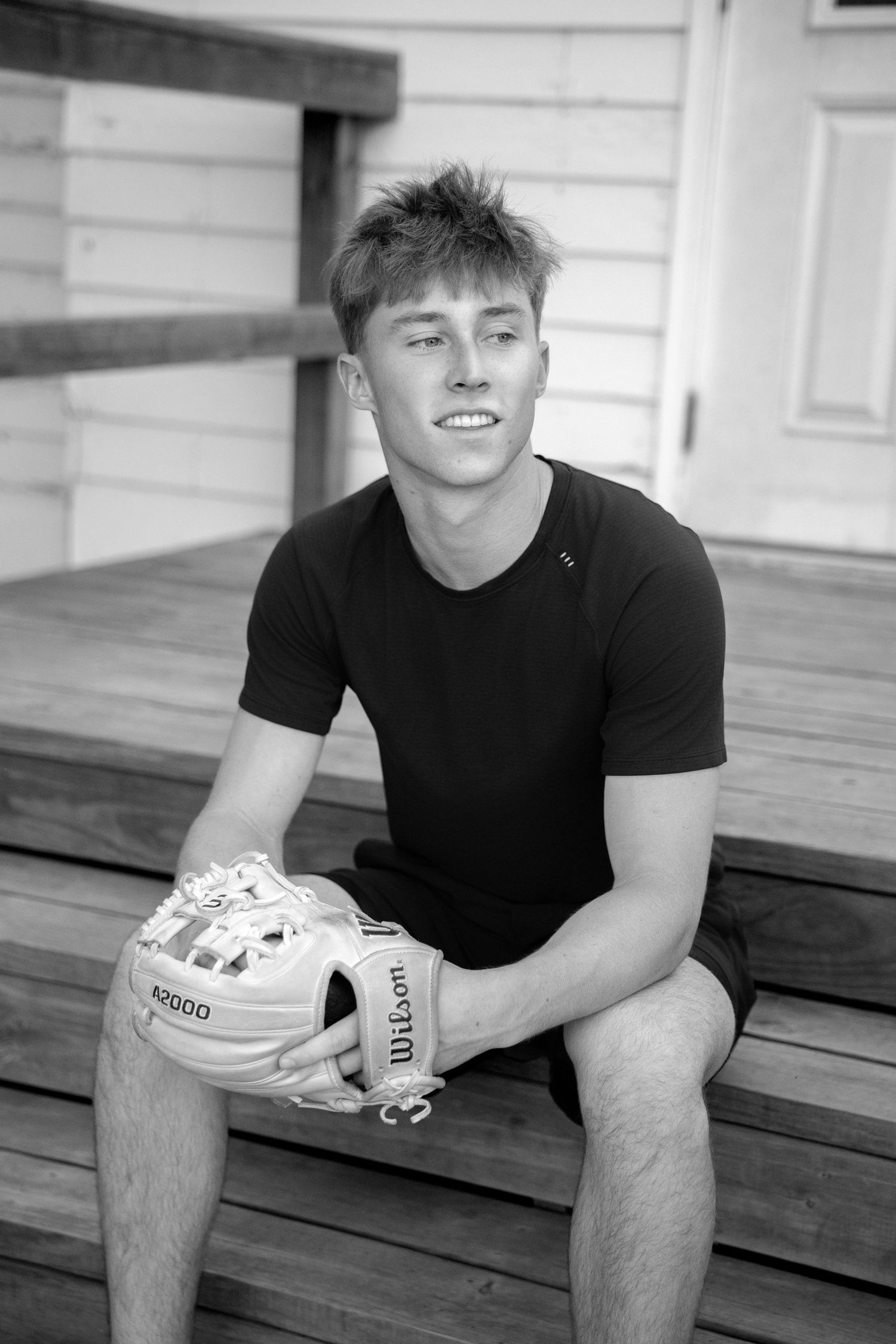 Young male athlete sits on wooden steps holding a Wilson A2000 baseball glove in a black t-shirt, black and white photo.