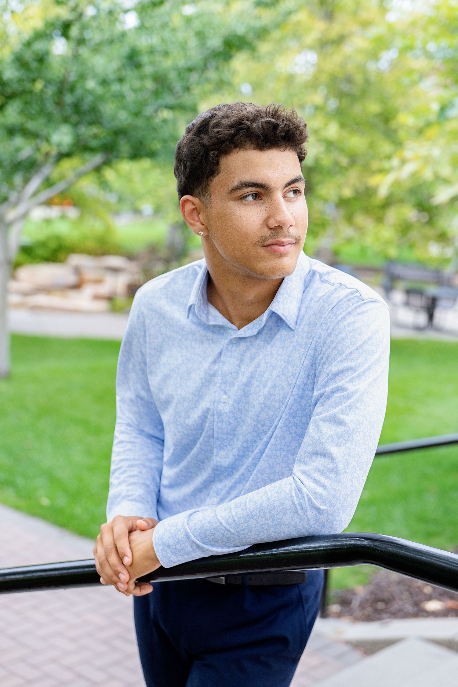 Young man in blue long-sleeve shirt leaning on railing in park.