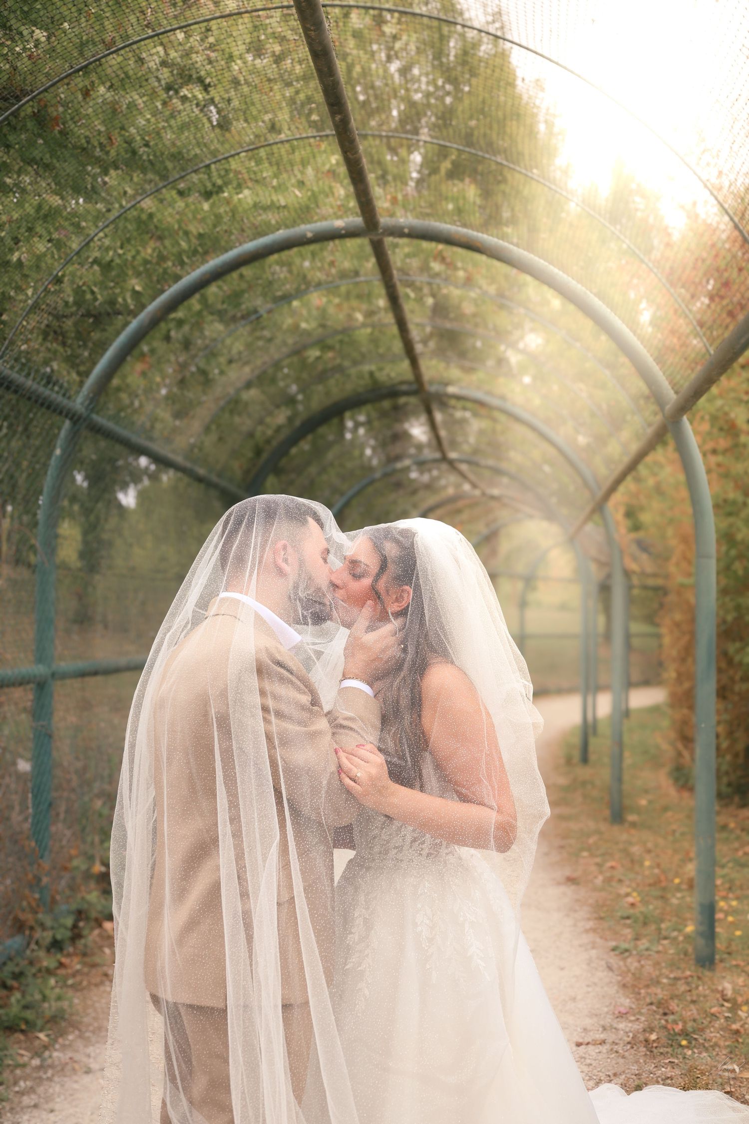 Bride and groom share a romantic kiss under a wedding veil in a sunlit garden archway.