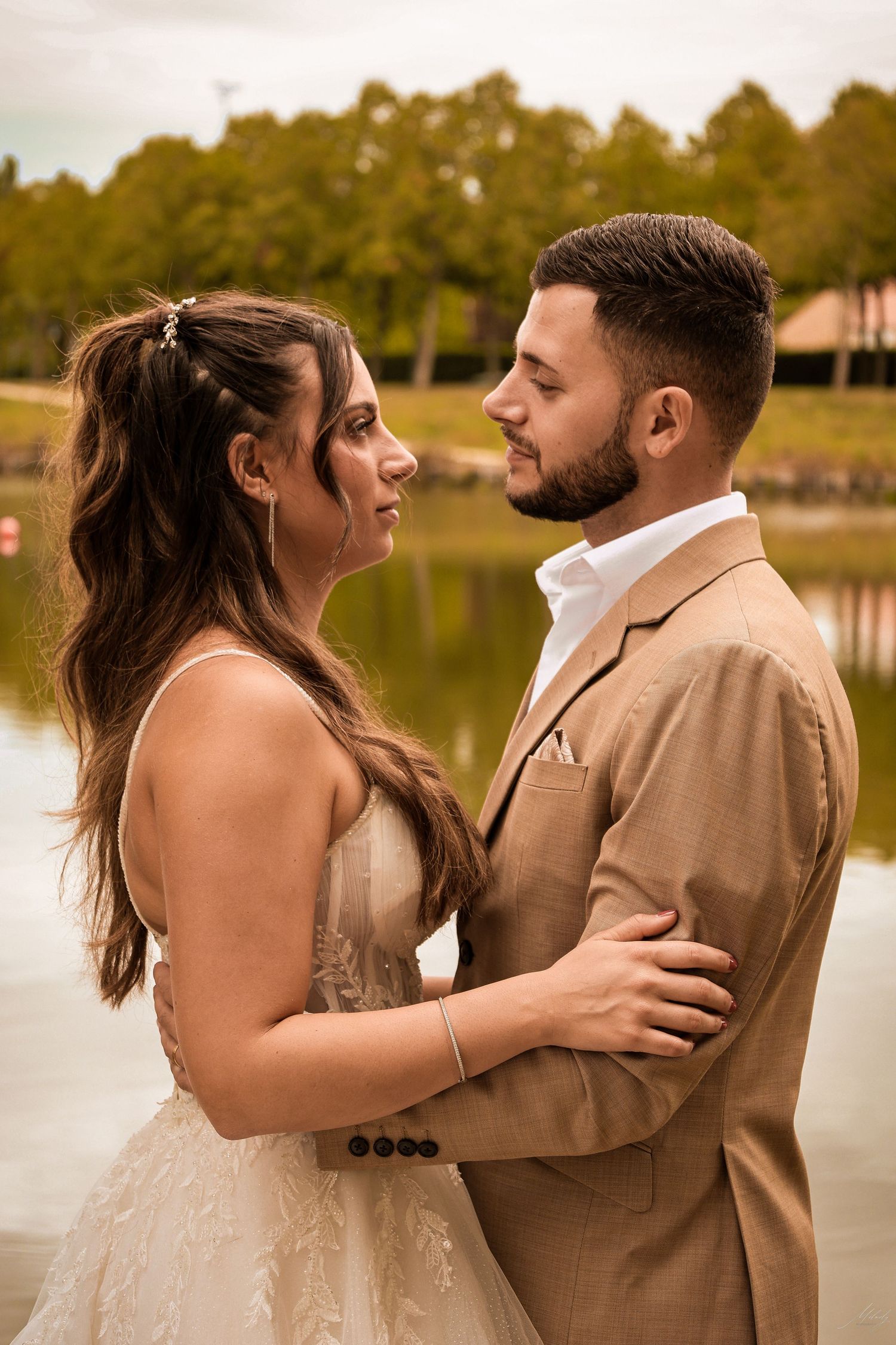 Bride and groom gazing at each other lovingly by a serene lake in outdoor wedding portrait.