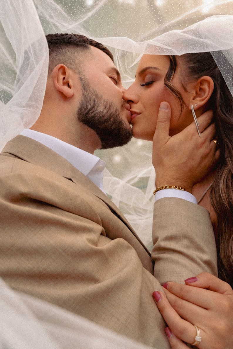 Couple sharing an intimate kiss beneath a sheer white bridal veil with sparkling light bokeh.