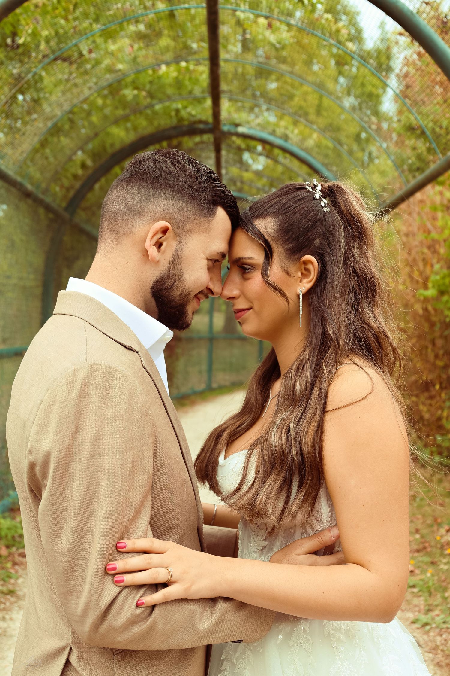 Newlywed couple touching foreheads romantically in a garden tunnel, bride in white lace gown.