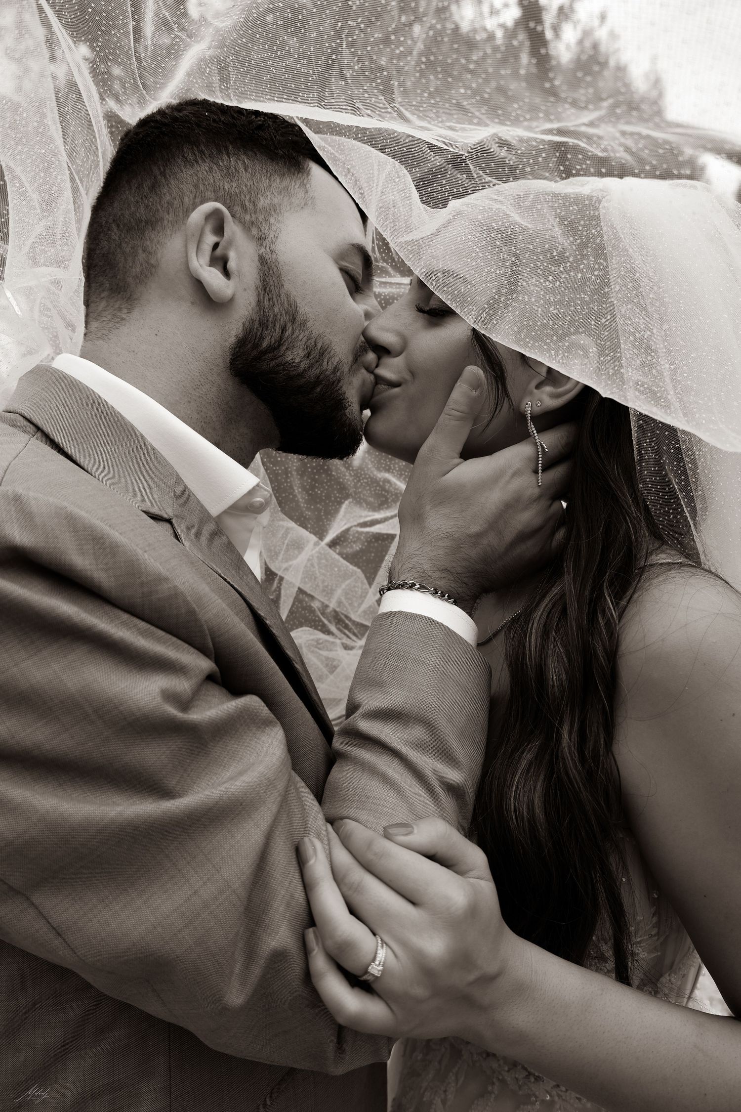 Romantic black and white photo of bride and groom kissing under a veil, groom cupping her face.